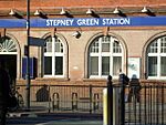 A red-bricked building with a rectangular, dark blue sign reading "STEPNEY GREEN STATION" in white letters and a man walking in front