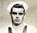 Head of a white man with dark, slicked-back hair wearing a white sailor's cap pushed up on his forehead and a sailor suit with a dark scarf tied around the neck.