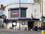 A grey-bricked building with a rectangular, dark blue sign reading "TOOTING BEC STATION" in white letters all under a blue sky with white clouds