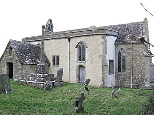 A small stone church seen from the south-east with a porch to the left, a protruding rendered south aisle in the middle, and a short chancel to the right.  On the west gable is a bellcote, and in front of the church is a churchyard cross
