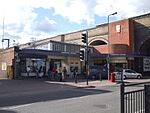A red-bricked building with a blue sign reading "GREENFORD STATION" in white letters and people in front all under a blue sky with white clouds