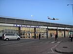 A dark grey building with a dark blue sign reading "HATTON CROSS STATION" in white letters all under a blue sky with an aeroplane flying through it