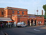 A red-bricked building with a blue sign reading "GOLDERS GREEN STATION" in white letters and people in front all under a blue sky with white clouds