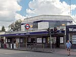 A grey building with a blue sign reading "EALING COMMON STATION" in white letters and two green trees in the background all under a blue sky