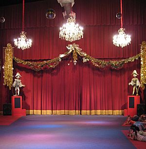 A stage flanked by large marionette puppets with large chandeliers hanging above