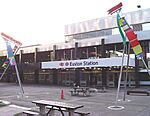 A white building with black columns and a white sign reading "EUSTON STATION" in blue letters and a traffic pilon in the foreground all under a white sky