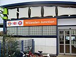 A white-panelled building with a rectangular, orange sign reading "Willesdon Junction" in white letters all under a light blue sky