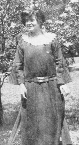 A young white woman with cropped hair, wearing a long loose-fitting dress with a white collar and a low-slung belt