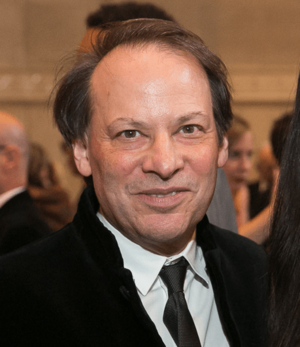 close-up of Adam Gopnik wearing a light striped shirt and dark blazer, with a headset microphone, looking intently just right of camera