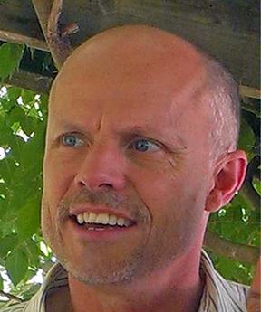 A bald white man with blue eyes, seen from an angle slightly below his face, with leaves in the back.