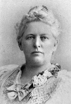 A white woman with white hair, wearing a light-colored dress with a neckline embellished with ribbons and flowers.