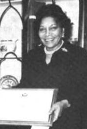 A black and white photograph of Barbara M. Watson, a Black woman, smiling, holding an award plaque. She is wearing a dark business suit and pearls. She is standing in front of a china cabinet.