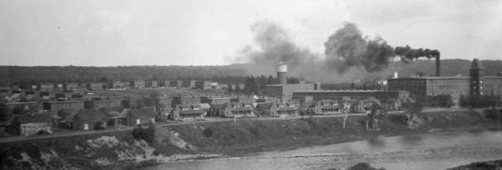 In the foreground is a river, with its far bank populated with many two-storey duplexes at left, and a large four-storey building to the right, behind which is a tall chimney releasing a black smoke billowing to the left.