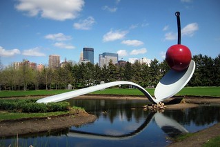 A large gray spoon straddles a shallow pond. On the tip of the spoon is a stylized red cherry with a black stem. The sculpture is surrounded by lawn and in the background, coniferous trees, behind which several skyscrapers stand. The sky is blue with several clouds.