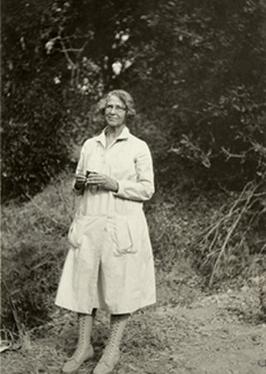Woman standing in front of plants, wearing a long coat and glasses