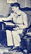A smiling woman in a short-sleeved shirt and pants sits on a chair with what appears to be a typewriter on her lap