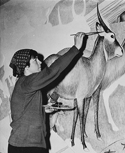Young woman in a kerchief reaches up to paint a detail of an antelope on a wall