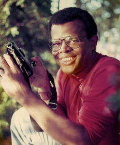A bespectacled, smiling man holds a camera