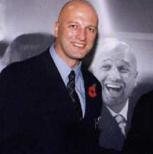 A bald man in a black suit posing in front of an earlier picture of himself.