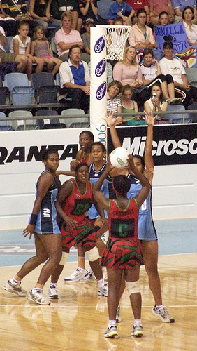 Six players in front of a netball basket. One is in the act of shooting, one is trying to block. Three are in red and three are in blue.