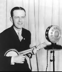 Photo of a young white man in 1920s outfit holding a banjo in front of a 1920s style radio microphone