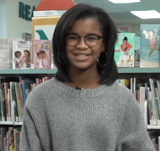 Marley Dias is in front of bookshelves at a library. She is wearing a gray sweater and silver hoop earrings. She has brown skin, straight black hair, and black-framed glasses.