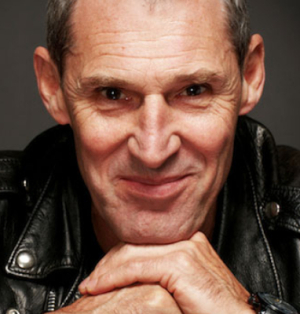 A man in a black leather jacket smiles at the camera, with his hands folded under his chin.