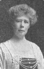 A middle-aged white woman with grey hair in a bouffant updo. She is wearing a dress with a square neckline and beaded embellishments across the bust.