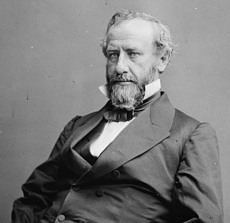 A man with receding, graying hair, a mustache, and a long beard wearing a dark jacket, vest, and tie and a white shirt