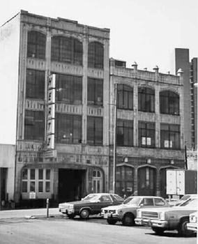 A black and white image of two commercial buildings, both three stories tall, the one on the left slightly higher, with large gently arched windows and some Gothic-style decorative touches. On the left is a vertical sign for Hertz rent-a-cars. There is a parking lot in the foreground with some vehicles