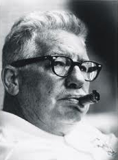 Candid black and white head-shot photograph of Rooney wearing a white shirt and black-rimmed eyeglasses chewing on an unlit cigar