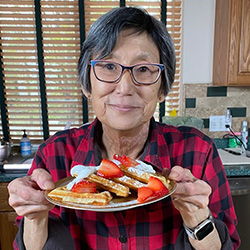A woman wearing glasses and a checked red and black shirt holds a plate of waffles with strawberries and whipped cream