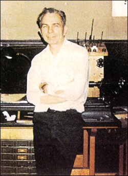 Color photograph of man leaning against a worktable with his arms crossed, a lathe, carpentry table and various tools visible behind him; he is approximately 55–60 years old with a greying head of hair and half-smile on his face.