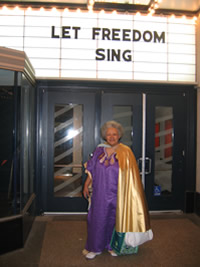 Herron is wearing a purple dress and gold cape, and standing outside a movie theater