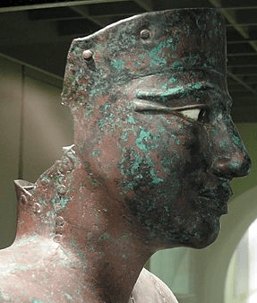 Closeup of the head of the statue of a man, made of greenish copper, its eyes inlaid in white and black stones