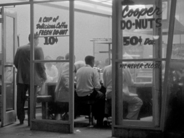 A black and white photograph of Cooper Donuts, with several men sitting with their back to glass doors. Text painted on the window reads "A cup of delicious coffee and fresh donut, 10 cents"
