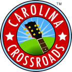 The Carolina Crossroads logo is circular with two concentric rings. The outer ring is red with the white text "Carolina" arcing around the top of the circle, "Crossroads" around the bottom, and five-pointed stars at the left and right of the ring. The inner circle has green in the bottom and blue in the top; the green appears to be a hill. The neck and headstock of a guitar rise from the bottom right edge; the headstock of the guitar is placed on the horizon formed between the green and blue sections. White streaks, evocative of sunbeams, radiate from the headstock.