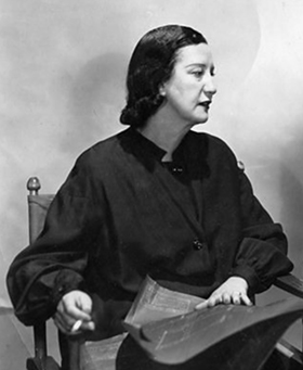 Black and white photograph of a women in a dark blouse, whose head is turned to the left, seated in a director's chair who has a cigarette in her right hand and whose left hand is resting on a blueprint in her lap.