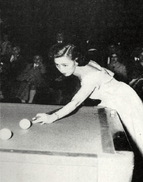 Black-and-white photo of Japanese woman in fancy dress, approximately 30 years old, stretched out over corner of carom billiards table with her cue stick in hand. Her eyes are intently focused on the shot in front of her, two billiards balls about a foot apart, with her cue tip about two inches behind the closest ball in position for the hit; the table scene is in spotlight and in the much darker background can be just made out spectators in chairs, all appearing to be men.