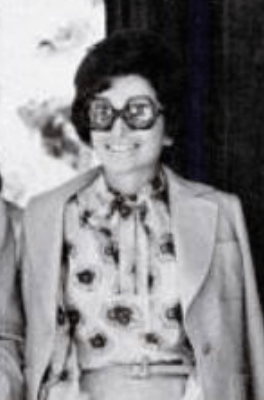 A smiling white woman with dark hair in a bouffant set, wearing a light-colored suit with a print blouse, and large sunglasses