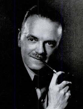 A middle-aged man with black hair and a moustache smokes a pipe