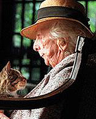 A color photograph of Marjory Stoneman Douglas late in her life. She is shown in profile, seated, with a cat on her lap. She is white-haired tanned and wrinkled. She wears a lapelled jacket and low-brimmed straw hat. She and the cat gaze at each other lovingly.