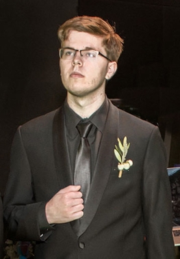Formally dressed young man in black tuxedo, black button down shirt, and black tie with glasses standing still