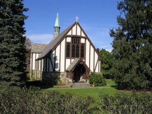 A cream-colored building with brown vertical beams on its front wall and a pointed roof with a cross on top. In the rear is a narrow green tower