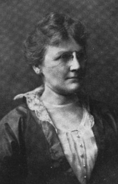 A white woman, wearing pince-nez eyeglasses and a lace-collared dress.