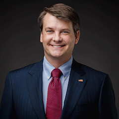 Official portrait of Luke Letlow smiling. He wears a navy blue suit, red tie, and an American flag lapel pin.