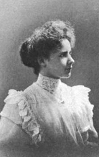 A young white woman wearing a white blouse with a high lace collar and pinafore-style ruffles. Her hair is in a large bun at the back of her head.