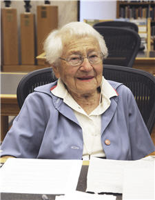 Photograph of Dr. Lucy Ozarin from 2012. She is smiling, while sitting at a desk with a few papers on the desk. She is wearing a white collared shirt and a blue jacket.
