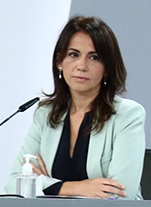 Silvia Calzón, wearing a light turquoise jacket over a black top, sits and looks slightly to the side in front of a black microphone and a pump-bottle of hand sanitizer.