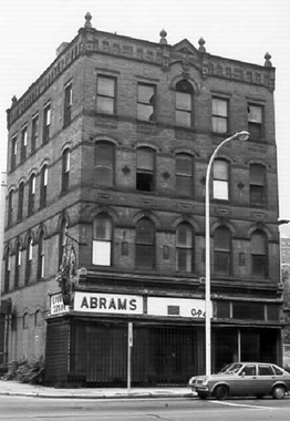 A black-and-white photograph of a four-story building with a flat ornamented roofline seen from across a city street and slightly to its right. On the ground floor storefront the word "Abrams" is prominent on the building's left.